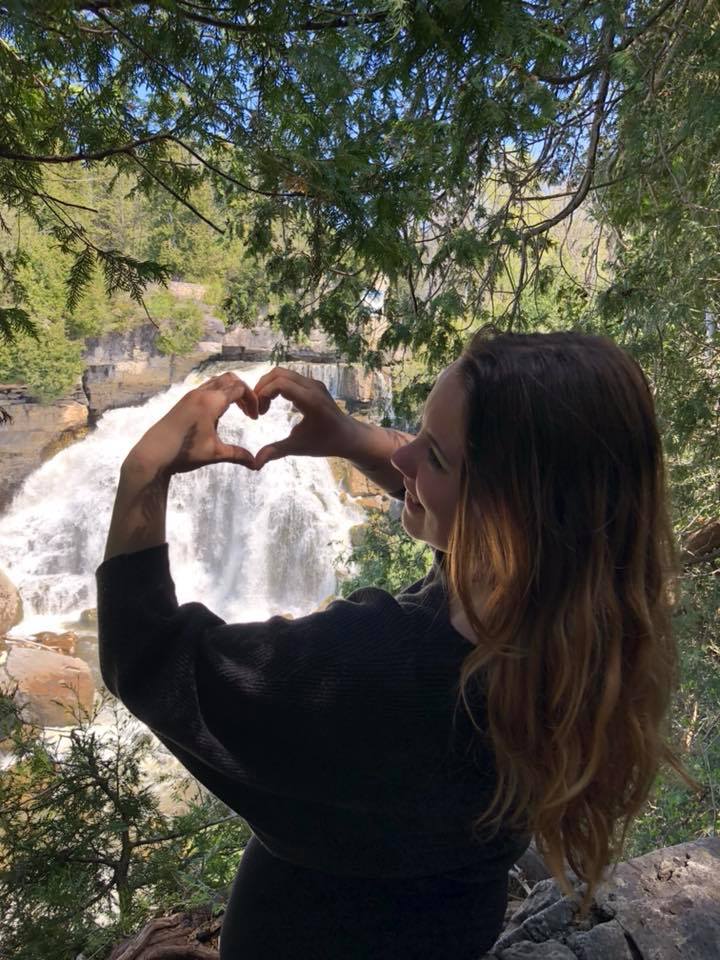 A woman making a heart with her hands in front of a waterfall